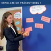 A woman in a suit is standing in front of a blue board with various notes and cloud-shaped cutouts. She is holding pink cards. Anteprima dell’immagine prodotto