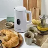 A white electric kettle is displayed on a table with croissants, tea cups, and lemons, suggesting a breakfast or tea time setting. Produktbild-Vorschau