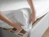 Hands are fitting a white sheet to a mattress on a bed frame. Produktbild-Vorschau