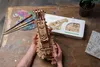 A person holding a wooden 3D puzzle of a hurdy-gurdy on a wooden table with art supplies and a plant. Produktbild-Vorschau