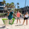 Three people playing a paddle ball game on a sandy beach with palm trees and buildings in the background. Produktbild-Vorschau