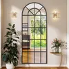 A large arched mirror with a black metal frame is shown in a room, reflecting a window with greenery. There is a plant on the left and a small table with flowers on the right. Produktbild-Vorschau