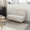 A cream-colored sofa bed with two pillows is placed on a light-colored rug in a room with a window and a black cabinet. Produktbild-Vorschau