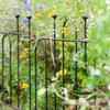 A black metal garden fence with decorative finials is shown in a garden setting, with colorful wildflowers and green foliage in the background. Podgląd zdjęcia produktu
