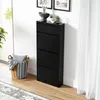 A black shoe cabinet with four flip drawers is placed against a white wall, next to a window with sheer curtains. A woven basket with a pillow and blanket sits on the floor beside the cabinet. Voir les images du produit