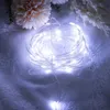 A coiled string of white LED lights on a textured white surface, with soft pink and white flowers in the background. Produktbild-Vorschau