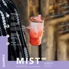 A person is holding a pink and orange gradient tumbler with a straw, with a blurred background of wooden structures. Podgląd zdjęcia produktu