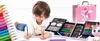 A young child is lying on the floor and drawing with colored pencils. There are several art supply cases open around the child, filled with various art supplies like colored pencils, crayons, and paints. A pink unicorn-themed art case is also visible in th Náhľad obrázku produktu