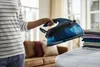 A person holding a steam iron over an ironing board, with folded clothes nearby. Náhľad obrázku produktu