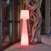 A tall, illuminated floor lamp with a conical base and a pleated shade stands on a wooden deck next to a large, gnarled tree trunk. Produktbild-Vorschau