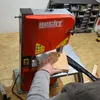 A person is using a red and silver bandsaw to cut a piece of wood. The background shows a workshop environment with storage bins filled with screws and hardware. Produktbild-Vorschau