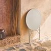A round, beige bistro table is folded and leaning against a textured wall, with a rustic wooden door and stone flooring. Produktbild-Vorschau