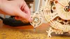 A hand is adjusting the pendulum of a wooden mechanical clock. Produktbild-Vorschau