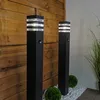 Two black bollard lights with clear glass sections are positioned in front of a wooden slatted wall, with small green bushes and white gravel on the ground. Produktbild-Vorschau