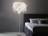 A floor lamp with a leaf-shaped lampshade stands next to a bed in a bedroom. Produktbild-Vorschau
