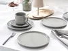 A table setting with grey porcelain dishes, a cup and saucer, cutlery, and a decorative tray with a lamp. Produktbild-Vorschau