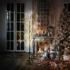 A decorative LED tree with warm white lights is placed in a cozy room with a fireplace and a Christmas tree in the background. Náhled obrázku produktu
