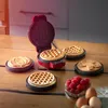 A red mini waffle maker is open on a wooden table, with waffles in various shapes in and around it. A bowl of raspberries and some eggs are in the foreground. Produktbild-Vorschau