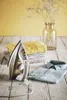 An iron and folded clothes are arranged on a wooden surface, with a vase of dried flowers in the background. Produktbild-Vorschau