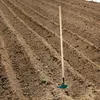 A garden hoe is standing upright in freshly tilled soil, with rows of soil visible. Produktbild-Vorschau