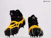 A pair of black hiking boots with yellow crampons and silver chains attached, displayed against a white background. Produktbild-Vorschau