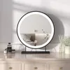 A round LED makeup mirror with a black frame sits on a wooden vanity table, reflecting a bedroom scene. Various cosmetic bottles and a diffuser are placed on the table. Voir les images du produit