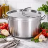 A large stainless steel pot with a lid, surrounded by fresh vegetables and a bowl of salt on a marble countertop. Produktbild-Vorschau