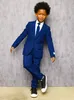 A young boy wearing a blue suit and sneakers, posing against a textured gray background. Anteprima dell’immagine prodotto