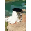 A black metal side table with a tray top sits on a stone patio next to a pool. A white towel is draped over the side of the table, and magazines are stored inside the wire basket. Produktbild-Vorschau