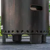 Close-up of the base of a dark-colored fire pit with a wood-grain texture, showing its legs and ventilation holes. Produktbild-Vorschau