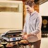 A man is using a contact grill to make sandwiches in a kitchen. Anteprima dell’immagine prodotto