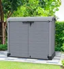 A grey plastic storage box with double doors sits on a paved patio area in a garden setting. Náhled obrázku produktu
