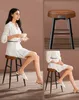 A woman is sitting on a bar stool, holding a cup and saucer. The image is split into multiple sections showing different views of the bar stool. Produktbild-Vorschau