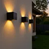 Three black cube-shaped outdoor wall lights mounted on a light-colored textured wall, casting warm yellow light downwards and upwards. The background shows a dark fence, green grass, and trees, suggesting an outdoor evening setting. Náhľad obrázku produktu