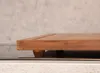 A close-up side view of a wooden cutting board with small wooden feet, resting on a reflective surface. Produktbild-Vorschau
