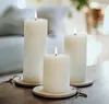 Three white pillar LED candles of varying heights are arranged on a wooden surface, with a blurred background of a room interior. Produktbild-Vorschau