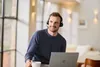 A man wearing a headset is working on a laptop in a bright room. Produktbild-Vorschau