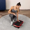 A woman is kneeling on a rug next to a vibration plate, holding a remote control. Náhled obrázku produktu
