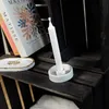 A close-up of a white candle in a white holder, placed on a dark wooden shelf next to books and a small decorative dish. Produktbild-Vorschau