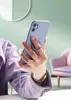 A person is holding a purple iPhone 13 Mini with a ring holder attached, taking a selfie. The background is a soft green with a decorative circular element and a pink armchair. Produktbild-Vorschau