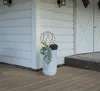 A decorative sphere with lights sits in a planter next to a white building with large doors. Produktbild-Vorschau