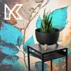 A black planter with a succulent plant on a black side table, set against a colorful, abstract floral background with a large white 'K' logo in the upper left corner. Produktbild-Vorschau