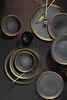 A collection of black and gold rimmed plates and bowls arranged on a dark textured surface. Produktbild-Vorschau