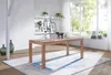 A wooden dining table is set in a bright, airy room with a large window overlooking greenery. Shelves with plants and pictures are visible on the wall. Anteprima dell’immagine prodotto