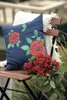 A navy blue pillow with a rose design sits on a wooden chair next to a bouquet of red roses. Produktbild-Vorschau