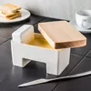 A butter dish with a wooden lid and a piece of butter inside, placed on a black tiled surface. Toasted bread and a cup of coffee are visible in the background. Podgląd zdjęcia produktu