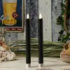 Two black LED candles with flickering warm white LED flames are displayed on a wooden surface, with a vintage beer advertisement poster in the background. Produktbild-Vorschau