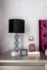 A digital alarm clock sits on a nightstand next to a lamp and a decorative jar. Náhľad obrázku produktu