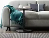A close-up of a C-shaped side table with a marble-like finish, placed next to a gray sofa. A teal throw blanket with fringe is draped over the sofa arm, and a book and cup are on the table. A striped rug is visible on the floor. Anteprima dell’immagine prodotto