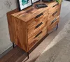 A wooden dresser with black handles is shown in a room with a concrete wall and wooden floor. Produktbild-Vorschau